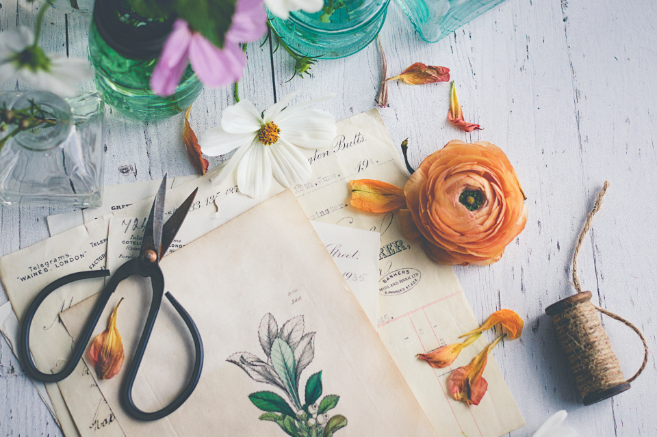 Botanical flatlay with flowers, scissors, paper, and glass jars on a white wood surface