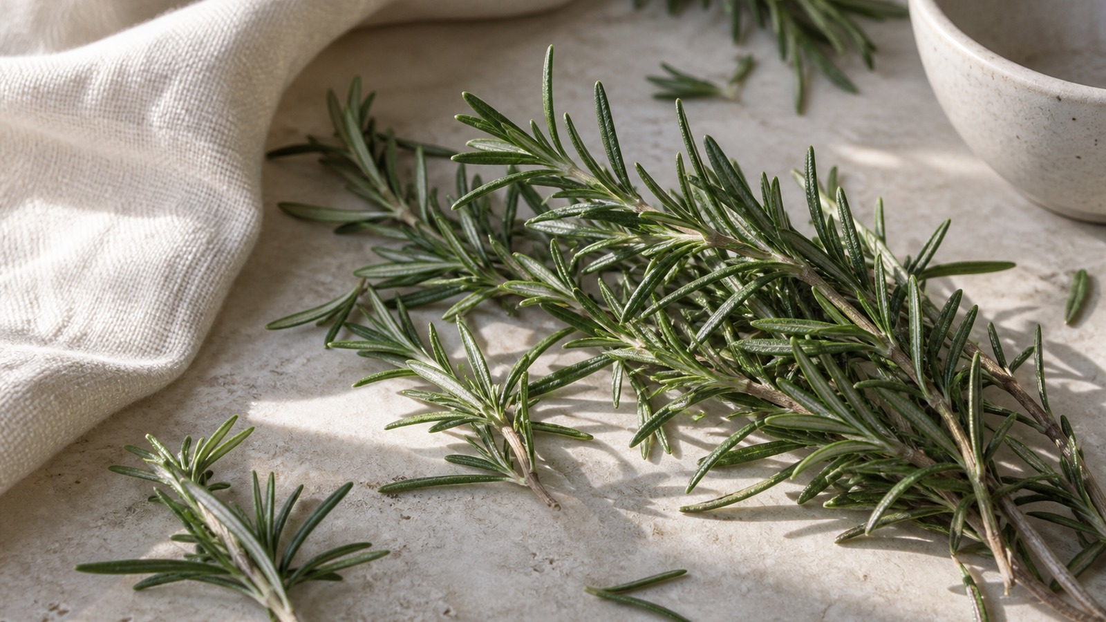Fresh rosemary sprigs on pale limestone beside ivory linen and an empty ceramic bowl