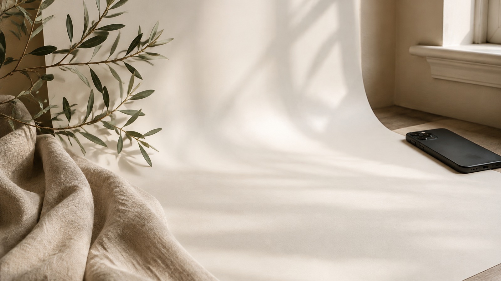 Phone beside an ivory photography backdrop, linen cloth, and olive branches in window light