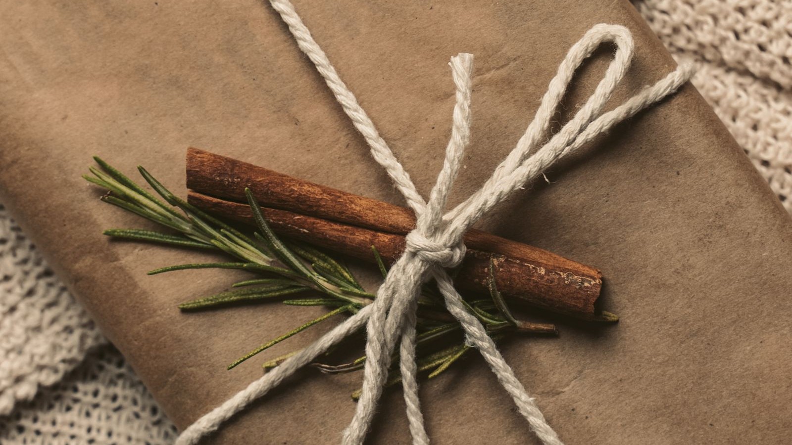 A kraft-paper gift tied with twine, rosemary, and cinnamon on a cream knitted blanket